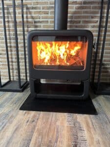 Poêle à bois moderne noir de marque DOVRE, rempli de flammes vives, devant un mur de briques rustiques et un sol en planches de bois clair.