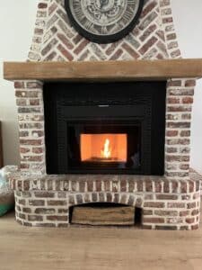 Insert de poêle à pellets Nobis allumé dans une cheminée en briques rouges à chevrons, surmontée d'une horloge et d'un manteau en bois.