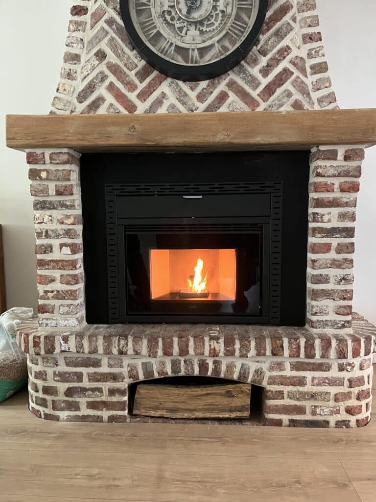 Insert de poêle à pellets Nobis allumé dans une cheminée en briques rouges à chevrons, surmontée d'une horloge et d'un manteau en bois.