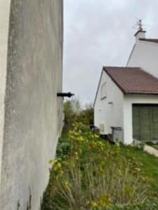 Émanation de vapeur blanche d'un conduit d'échappement noir fixé au mur extérieur d'un bâtiment, près d'une zone végétalisée et d'une autre maison.