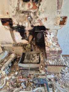 Démolition d'une cheminée encastrée montrant des dommages importants dus à la suie noire, des briques exposées, une grille de foyer métallique et des débris de démolition avec un marteau-perforateur Bosch.