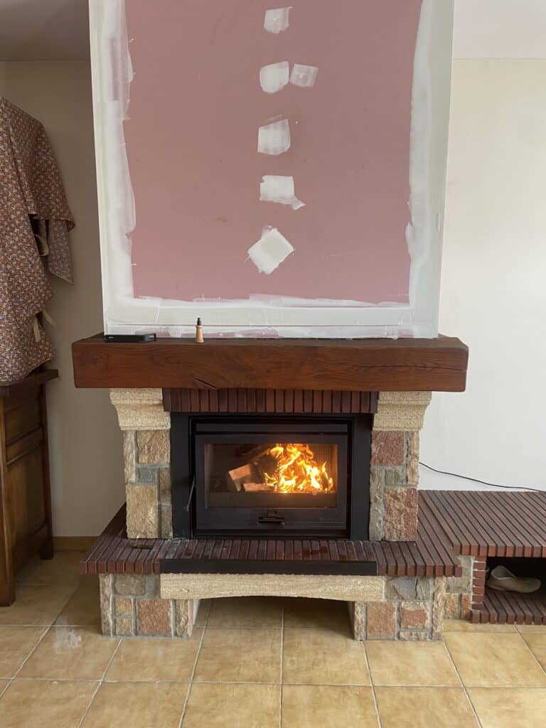 Cheminée avec insert en fonte allumé, manteau en bois et entourage en pierre, mur rose en cours de rénovation avec enduit blanc.