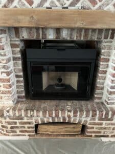 Insert de cheminée noir Nobis installé dans un foyer en briques rouges rustiques avec manteau de bois brut et bûches stockées en dessous.