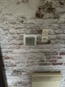 Mur de briques anciennes peint en blanc avec peinture écaillée. Ventilation carrée, interrupteur et prise électrique européenne crème visibles.