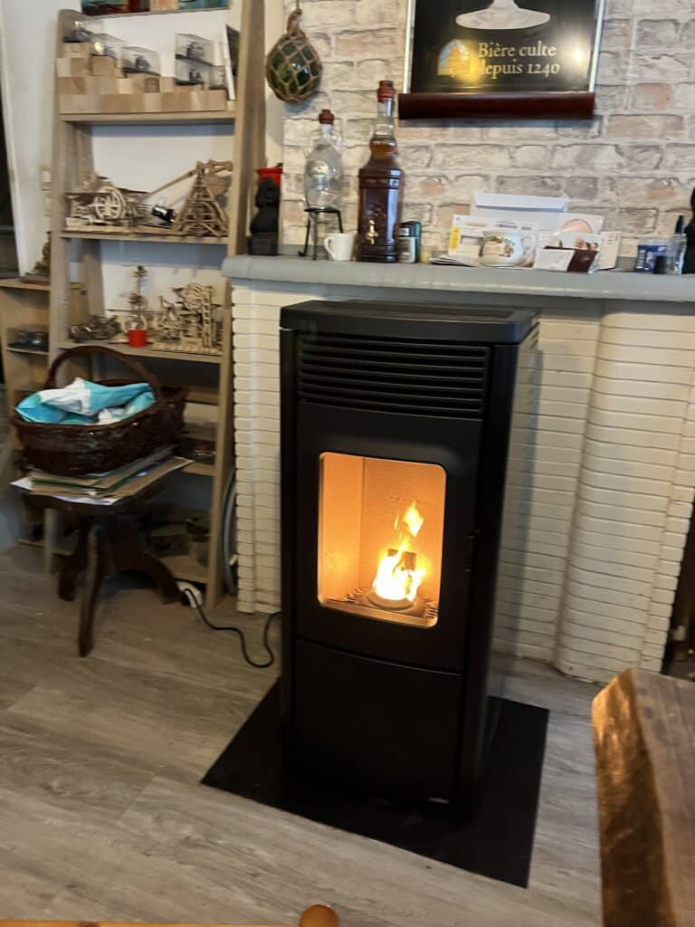 Poêle à granulés noir allumé devant une cheminée avec une inscription "Bière culte depuis 1240", à côté d'étagères remplies de maquettes en bois.