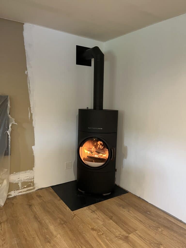 Poêle à bois Austroflamm noir allumé avec feu vif, installé sur un socle noir, dans une pièce en rénovation avec murs en cours de peinture.