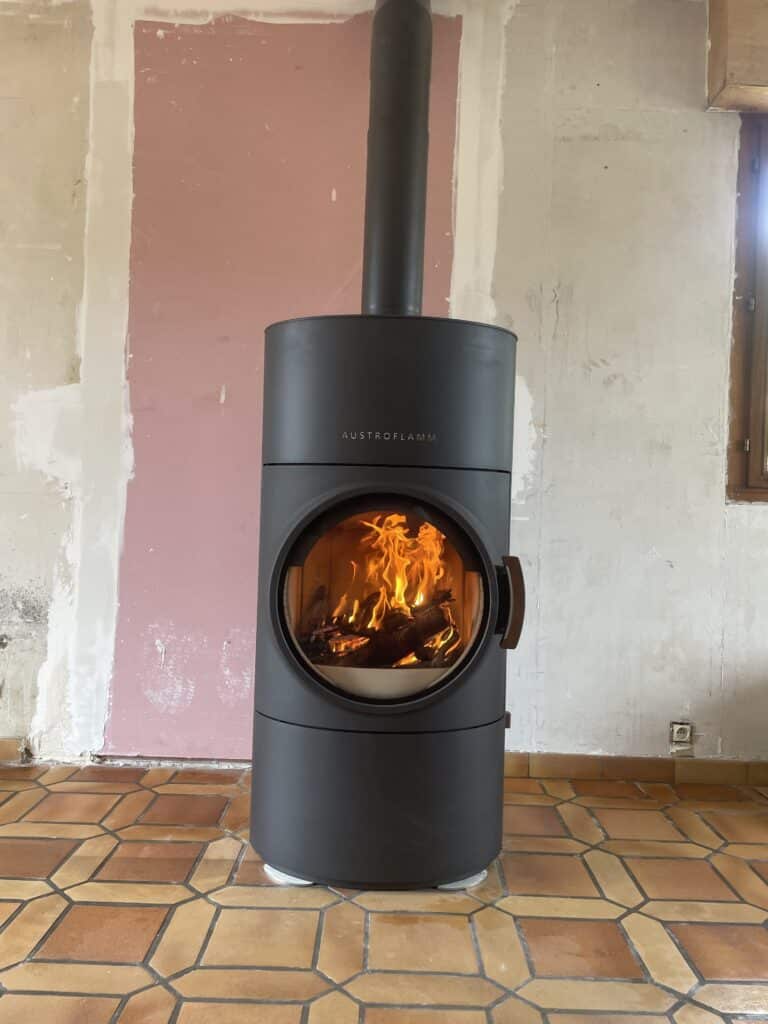 Poêle à bois Austroflamm noir et cylindrique en fonctionnement avec flammes vives, installé sur un sol carrelé ocre dans une pièce en rénovation.