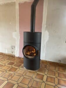 Poêle à bois Austroflamm noir en fonctionnement, avec flammes visibles, dans une pièce en rénovation avec murs bruts et carrelage terre cuite.