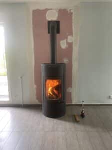 Poêle à bois noir allumé dans une pièce en cours de rénovation, murs à peindre, sol carrelé gris.