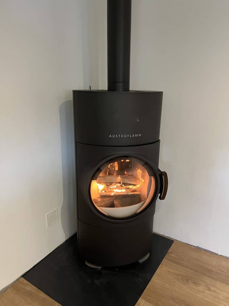 Poêle à bois cylindrique noir Austroflamm avec feu vif, porte ronde et conduit d'évacuation, posé sur une plaque noire près d'un mur blanc.