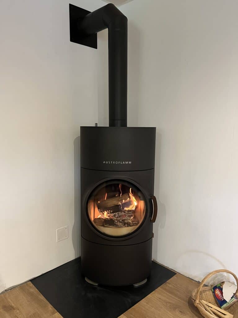 Poêle à bois moderne AUSTROFLAMM noir dans un coin, flammes visibles à travers la porte ronde, raccordement noir au mur.