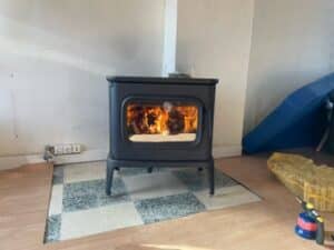 Poêle à bois noir en fonte allumé, dégageant une chaleur agréable, posé sur un tapis quadrillé devant un mur clair.