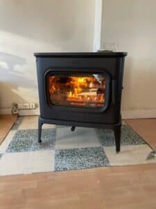 Poêle à bois noir moderne avec vitre, flammes vives et bûches ardentes, posé sur un tapis à carreaux devant une prise murale.