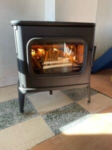 Poêle à bois moderne en fonte noir, vitré, avec bûches incandescentes à l'intérieur, posé sur un sol carrelé clair et foncé.