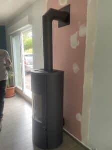 Poêle à bois noir cylindrique avec conduit d'évacuation coudé, installation en cours près d'une porte-fenêtre.