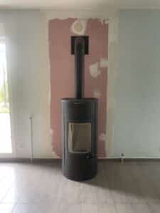 Poêle à bois noir installé devant un mur en rénovation avec enduit rose et trous de ventilation visibles.