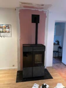 Poêle à bois moderne noir installé devant un mur en cours de rénovation (plâtre rose visible), avec cheminée, notices 'nobis' et télécommandes au sol.