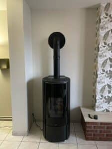 Poêle à bois moderne noir et cylindrique installé contre un mur clair, avec conduit d'évacuation vertical noir sortant par le haut et raccordé au mur.