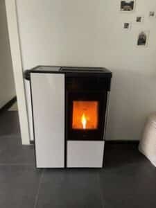 Poêle à granulés moderne, blanc et noir, avec flamme visible, installé sur sol carrelé sombre, près d'un mur blanc décoré de petites photos.