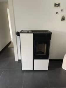 Poêle à granulés moderne blanc et noir, posé sur un sol carrelé foncé, avec un conduit d'évacuation visible à gauche.