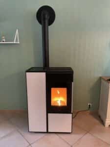 Poêle à granulés moderne blanc et noir allumé, avec une sortie de fumée noire verticale, installé dans une pièce aux murs vert d'eau.