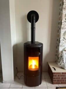 Poêle à granulés moderne, marron foncé, en fonctionnement avec flamme visible, raccordé à un conduit d'évacuation noir vertical contre un mur clair.