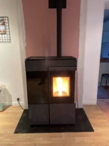 Poêle à granulés moderne noir allumé, posé sur un socle noir devant un mur en cours de rénovation, partiellement peint en rose saumon.
