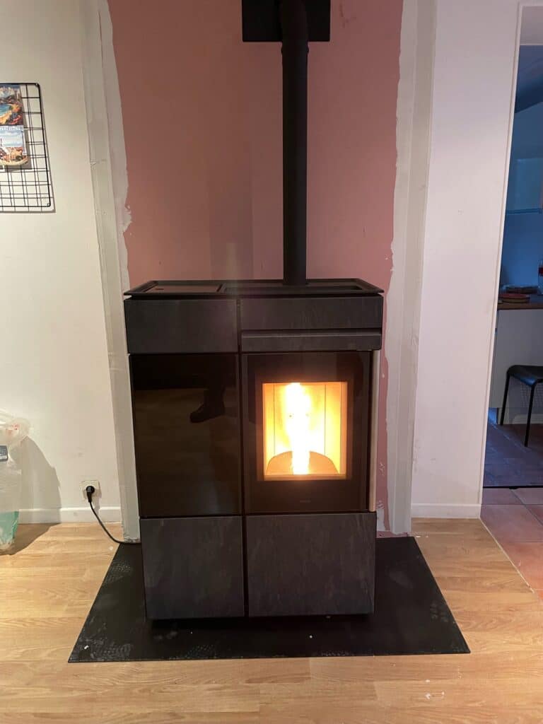 Poêle à granulés moderne noir allumé, posé sur un socle noir devant un mur en cours de rénovation, partiellement peint en rose saumon.