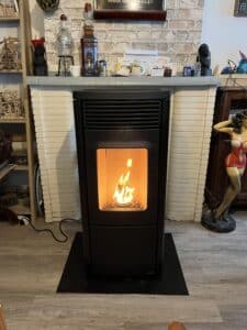 Poêle à granulés noir Nobis en marche avec flammes vives, devant une cheminée en briques claires, près d'une figurine de femme pin-up rouge.