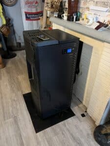 Poêle à granulés noir avec afficheur numérique, posé sur un tapis noir devant un manteau de cheminée décoré d'objets, dont une tasse "SOUP" et des lettres.