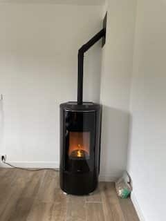 Poêle à granulés noir, moderne, cylindrique, allumé, avec conduit d'évacuation noir montant au mur blanc, sur plancher imitation bois.