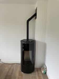 Poêle à granulés noir, moderne et cylindrique, raccordé à un conduit d'évacuation noir montant verticalement puis coudé vers le mur blanc. Sac de granulés au sol.