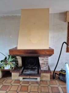 Panneau de bois brun clair, peut-être MDF, est installé devant la hotte d'un barbecue ou d'une cheminée en briques et bois, en rénovation.