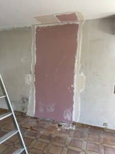 Mur en cours de rénovation avec plaque de plâtre rose visible, joints enduits, traces noires au sol et échelle en aluminium à gauche.