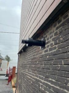 Ventouse d'évacuation de chaudière noire fixée sur un mur de briques sombres, sous un revêtement de façade brun clair.
