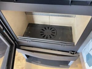 Foyer ouvert d'un insert de cheminée moderne. Parois en chamotte beige, grille en fonte noire, rampe de bûches et barre d'air secondaire.