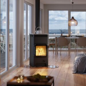 Poêle à bois noir AUSTROFLAMM avec feu dans un intérieur moderne et chaleureux. Grande baie vitrée et plancher en bois clair face à la mer.