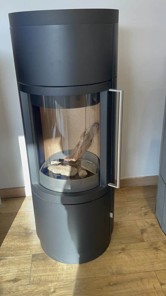 Poêle à bois cylindrique moderne noir mat | Design Hase Poêle à bois moderne cylindrique noir mat (marque hase), porte vitrée, poignée acier brossé. Bûches sur plancher chêne clair.
