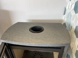 Plateau gris moucheté d'un insert de poêle à bois, montrant le raccord du conduit de fumée noir. Décor mural carrelé 3D.