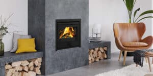 Salon moderne avec foyer encastré noir et bois de chauffage visible. Mur gris anthracite texturé. Fauteuil en cuir marron et coussin jaune vif.