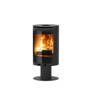 Poêle à bois moderne noir sur pied, de forme cylindrique, avec large vitre incurvée montrant de vives flammes de bois et un logo JM.