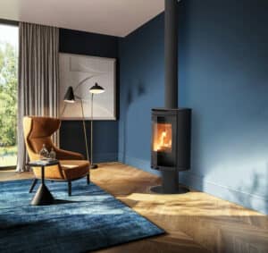 Poêle à bois noir et fauteuil orange vif dans un salon moderne aux murs bleus. Sol en parquet chevron et grand tapis bleu.